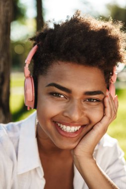 Olumlu neşe saçan Afrikalı kadının fotoğrafı. Parkta çimlerin üzerinde oturmuş kulaklıkla müzik dinliyor.