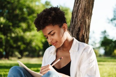 Konsantre olmuş Afrikalı bir kadının fotoğrafı. Parkta çimlerin üzerinde oturmuş deftere veya günlüğe notlar yazarken.