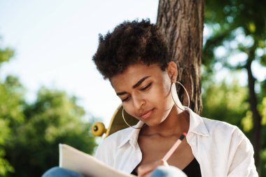 Çekici, gülümseyen, Afro-Afrikalı bir kadın parkta günlük tutuyor.