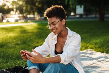 İyimser memnun Afrikalı bir kadının çimlerin üzerinde oturduğu ve cep telefonu kullandığı bir fotoğraf.