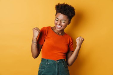 Heyecanlı bir Afrikalı Amerikalı kadının gülümsemesi ve kazanan jestinin izole edilmiş sarı arka plan görüntüsü