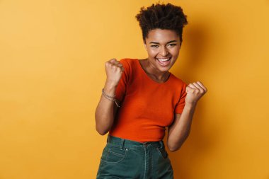 Heyecanlı bir Afrikalı Amerikalı kadının gülümsemesi ve kazanan jestinin izole edilmiş sarı arka plan görüntüsü
