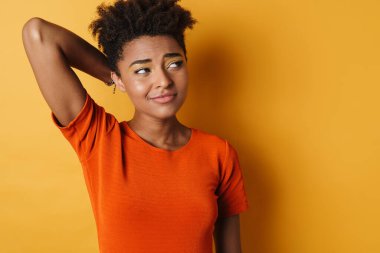 Kafası karışmış Afrikalı Amerikalı bir kadının, izole edilmiş sarı arka plana bakıp poz vermesi.