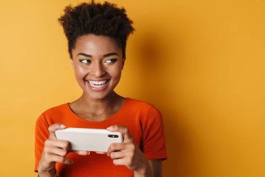 Neşeli Afro-Amerikan kadın görüntüsü cep telefonundan izole edilmiş sarı arka planda oyun oynuyor