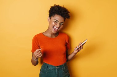 Heyecanlı Afrikalı Amerikalı bir kadının kazanan jesti ve cep telefonu sarı arka planda.