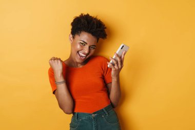 Heyecanlı Afrikalı Amerikalı bir kadının kazanan jesti ve cep telefonu sarı arka planda.