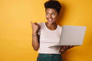 Afro-Amerikalı neşeli bir kadının parmak göstermesi ve dizüstü bilgisayarla izole edilmiş sarı arka plan kullanması