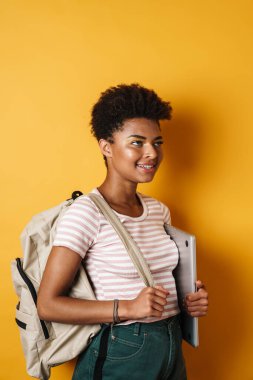 Neşeli Afrikalı Amerikalı bir kadının elinde dizüstü bilgisayarla gülümserken izole edilmiş sarı bir arka plan.