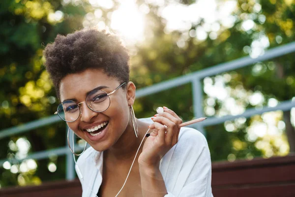 Parkta oturmuş müzik dinleyen neşeli, mutlu bir Afrikalı kadının fotoğrafı.
