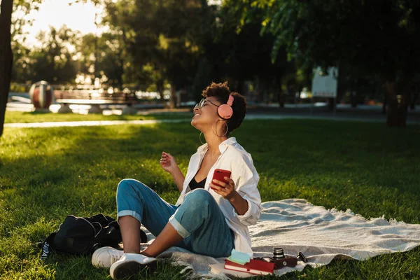 Neşeli, neşeli Afrikalı bir kadının çimlerin üzerinde oturmuş kulaklıkla müzik dinlerken ve cep telefonu kullanırken fotoğrafı.