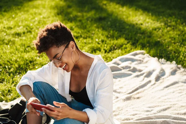 Fotoğrafta gülümseyen iyimser Afrikalı kadın çimlerin üzerinde parkta oturuyor ve cep telefonu kullanıyor.