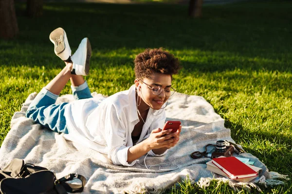 Fotoğrafta gülümseyen Afro-Afrikalı bir kadın çimlerin üzerinde uzanıyor ve müzik dinlerken cep telefonu kullanıyor.