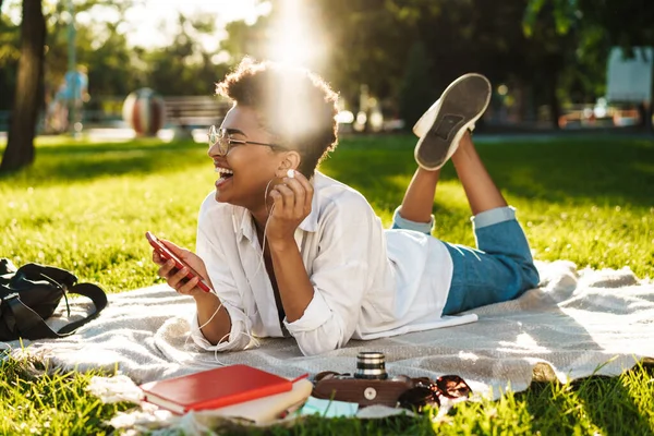 Neşeli Afrikalı bir kadının çimlerin üzerinde uzanıp müzik dinlerken cep telefonu kullanması.