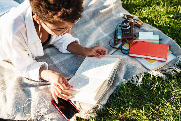 Изображение сконцентрированной африканской женщины, лежащей в парке на траве и читающей книгу
