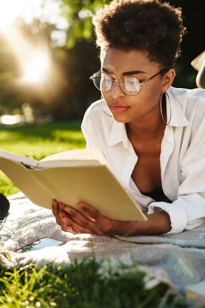 Ciddi bir Afrikalı kadının fotoğrafı. Parkta çimlerin üzerinde uzanmış kitap okuyor.