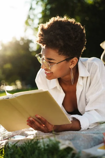 Mutlu, gülümseyen Afrikalı bir kadının çimlerin üzerinde uzanmış kitap okuduğu görüntüsü.