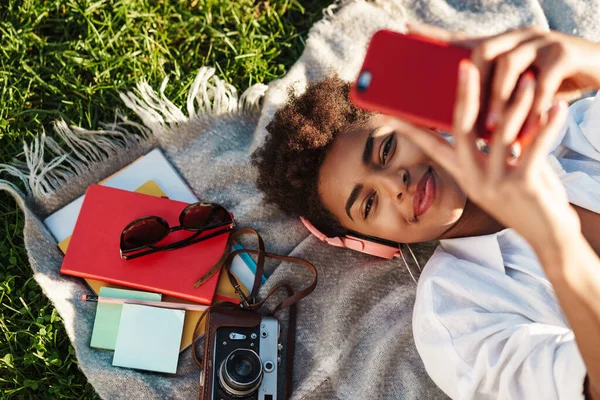 Gülümseyen Afrikalı bir kadının çimlerin üzerinde uzanmış cep telefonuyla müzik dinlediği bir görüntü.