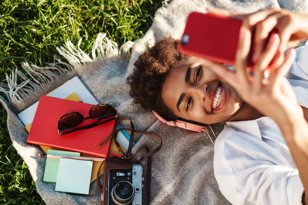 Mutlu, neşeli, Afrikalı bir kadının çimlerin üzerinde uzanıp cep telefonu kullanırken müzik dinlediği görüntüsü.