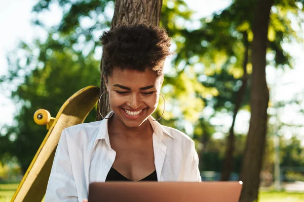 Mutlu, neşeli Afrikalı bir kadının çimlerin üzerinde oturmuş dizüstü bilgisayar kullanması.