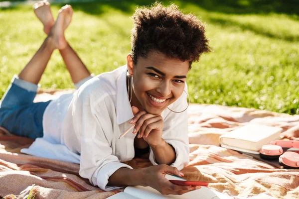 Neşeli gülümseyen Afrikalı bir kadının fotoğrafı dışarıda çimlerin üzerinde uzanıyor ve cep telefonu kullanıyor.