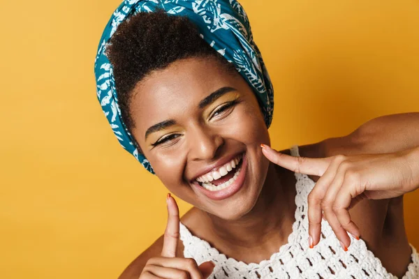 Image of happy african american woman laughing and pointing fingers at her cheeks isolated yellow background