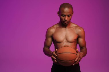Basketbolla poz veren yarı çıplak Afro-Amerikan bir adamın görüntüsü mor duvarın üzerinde izole edilmiş.