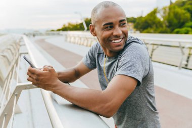 Sportif, genç Afrikalı bir adam açık havada dinlenirken, köprüde beklerken akıllı bir telefonla daktilo yazıyor.