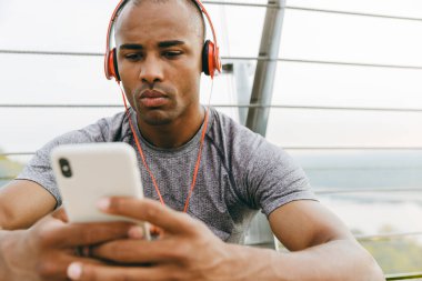 Rahat Afrikalı sporcu köprüde duruyor, kulaklık takıyor, cep telefonu kullanıyor.