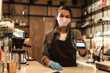 Tıbbi yüz maskesi takan kadın tezgahtaki siparişin parasını ödemek için elinde nakitsiz terminal tutuyor.