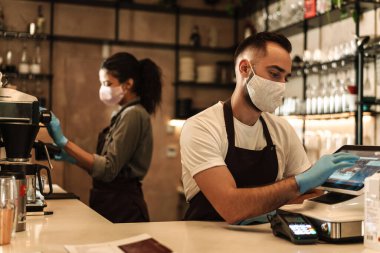 İçeride karantina sırasında resepsiyonda kahve servisi yapan tıbbi maske takan iki barmen.