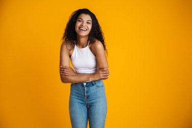 Çekici, mutlu, sıradan kıyafetler giyen Afrikalı bir kadın sarı arka planda tek başına duruyor.