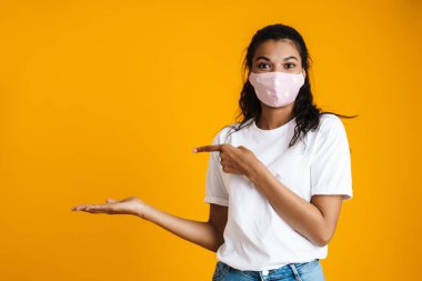 Tıbbi maske takan güzel, mutlu genç bir kadın, sarı arka planı işaret ediyor.