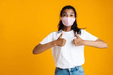 Sarı arka planda beliren tıbbi maske takan mutlu, genç Afrikalı kadın.