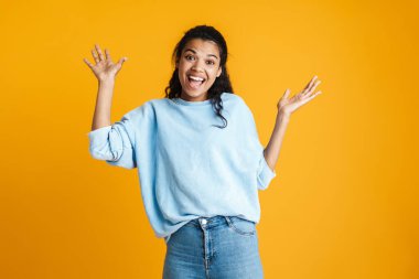 Çekici, mutlu, günlük kıyafetler giyen Afrikalı bir kadın sarı arka planda tek başına duruyor ve başarıyı kutluyor.