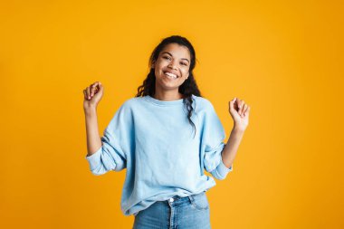 Çekici, mutlu, günlük kıyafetler giyen Afrikalı bir kadın sarı arka planda tek başına duruyor ve başarıyı kutluyor.