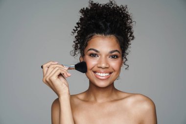 Tişörtsüz Afro-Amerikalı bir kadının toz fırçası kullanması ve gri duvarda sırıtışı.