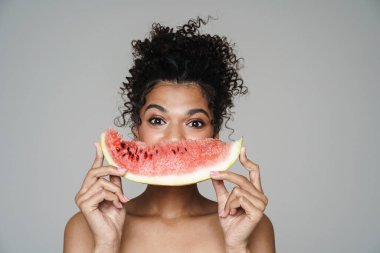 Gri duvarda izole edilmiş karpuzla poz veren üstsüz Afro-Amerikan kadını.