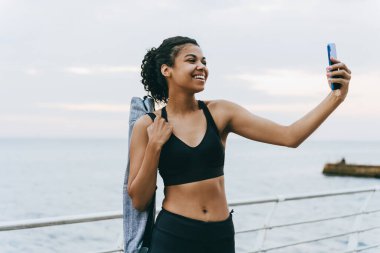 Gülümseyen Afro-Amerikan sporcu kadının gezinti alanında cep telefonuyla selfie çekişi.