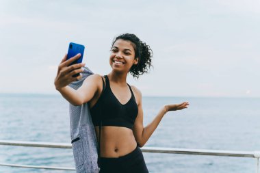 Gülümseyen Afrikalı Amerikalı sporcu kadının cep telefonuyla selfie çekerken ve gezinti alanında fotokopi çekerken görüntüsü