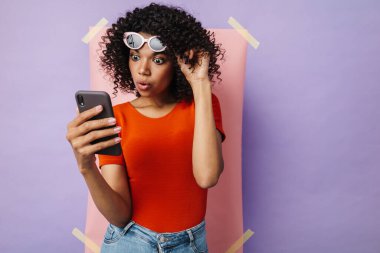 Çok renkli arka planda izole edilmiş cep telefonu kullanan şaşırmış Afrikalı Amerikalı kadın görüntüsü