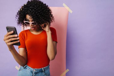 Çok renkli arka planda izole edilmiş cep telefonu kullanan şaşırmış Afrikalı Amerikalı kadın görüntüsü