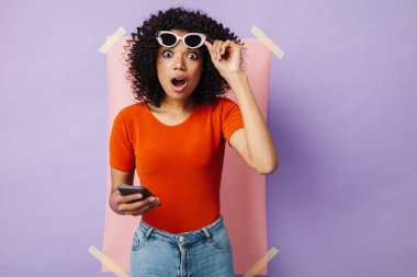 Çok renkli arka planda izole edilmiş cep telefonu kullanan şaşırmış Afrikalı Amerikalı kadın görüntüsü