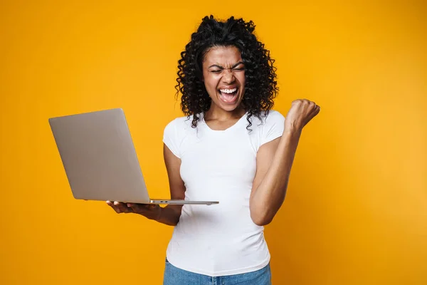 Image of excited african american woman making winner gesture and using laptop isolated over yellow background