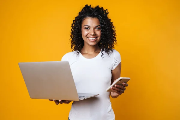 Image of happy african american woman holding cellphone and laptop isolated over yellow background