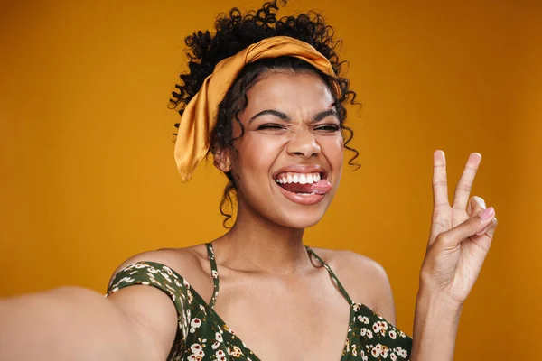黄色の壁の上に隔離された自撮りをしながら平和サインを示す幸せなアフリカ系アメリカ人女性のイメージ — ストック写真