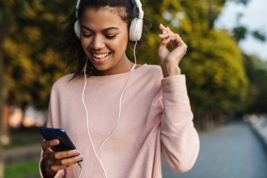 Yazlık parkta dans ederken cep telefonu ve kulaklık kullanan gülümseyen Afrikalı Amerikalı kız.