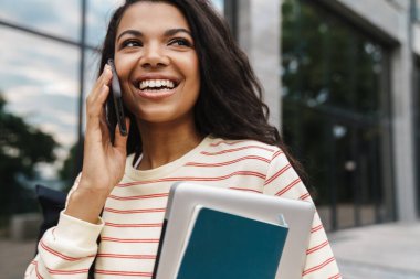 Gülümseyen Afrikalı Amerikalı bir kızın şehir caddesinde dizüstü bilgisayarla yürürken cep telefonuyla konuşması.