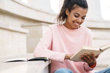 Dışarıda merdivenlerde oturmuş kitap okuyan neşeli Afro-Amerikan öğrenci resmi.