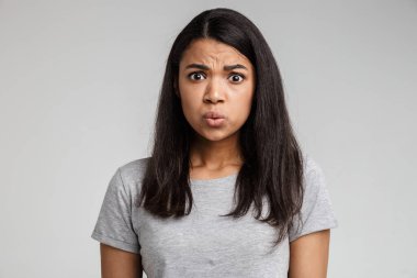 Kafası karışmış, günlük kıyafetler giymiş, gri arka planda tek başına duran Afrikalı bir kadın.
