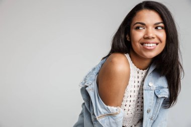 Uzun siyah saçlı, mutlu ve gülümseyen Afrikalı bir kadının portresi. Gri arka planda tek başına dikilip gözlerini kaçırıyor.